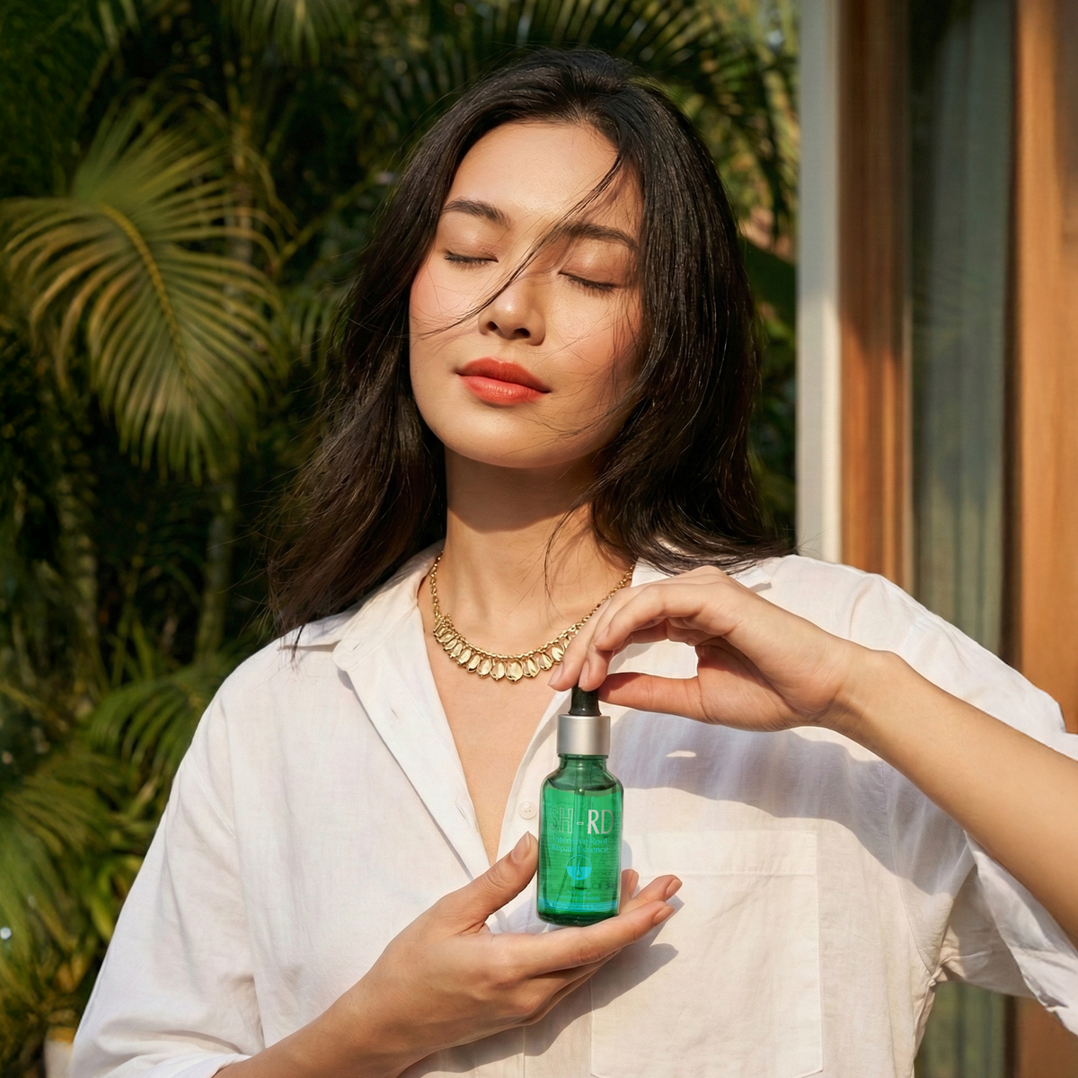 A woman in a white button-up shirt holds a small green bottle with a silver dropper against a warm, sunlit background with palm leaves.