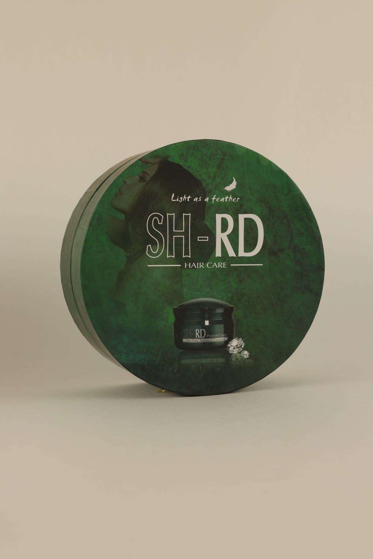A round green SH-RD Hair Care tin with a dark green textured design, featuring white text and a small black jar illustration, set against a solid beige background.