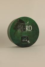 A round green SH-RD Hair Care tin with a dark green textured design, featuring white text and a small black jar illustration, set against a solid beige background.