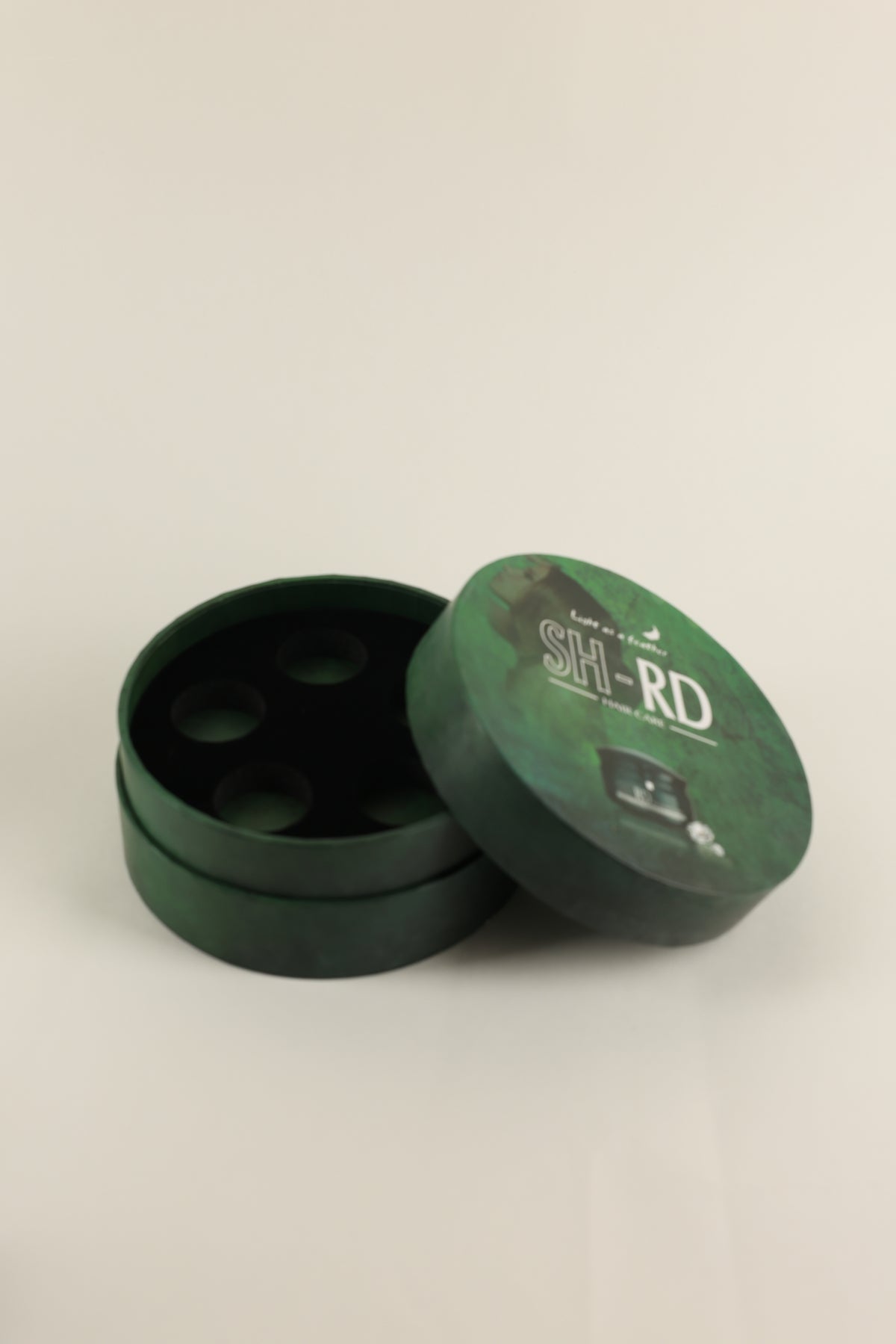 A round dark green cylindrical container with its lid leaning against it, shown on a plain light background. The lid has white printed text reading 'SH-RD'.