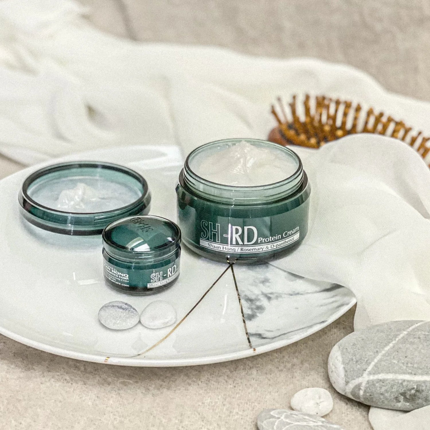 Three jars of SH-IRD Protein Cream on a marble tray with a wooden comb and white fabric in the background.