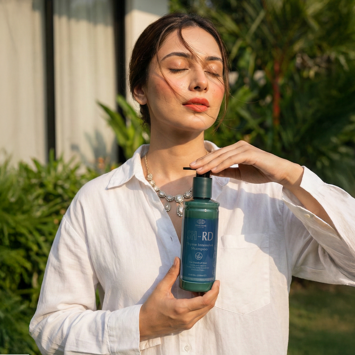 A woman in a white button-up shirt holds and dispenses liquid from a dark teal bottle labeled 'DERMA-RD' in an outdoor garden setting.
