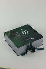 A rectangular gift box with a dark green textured lid and silver-gray sides, tied with a dark green ribbon. The lid features white text reading 'SH RD' and a small product photo, photographed on a light gray background.