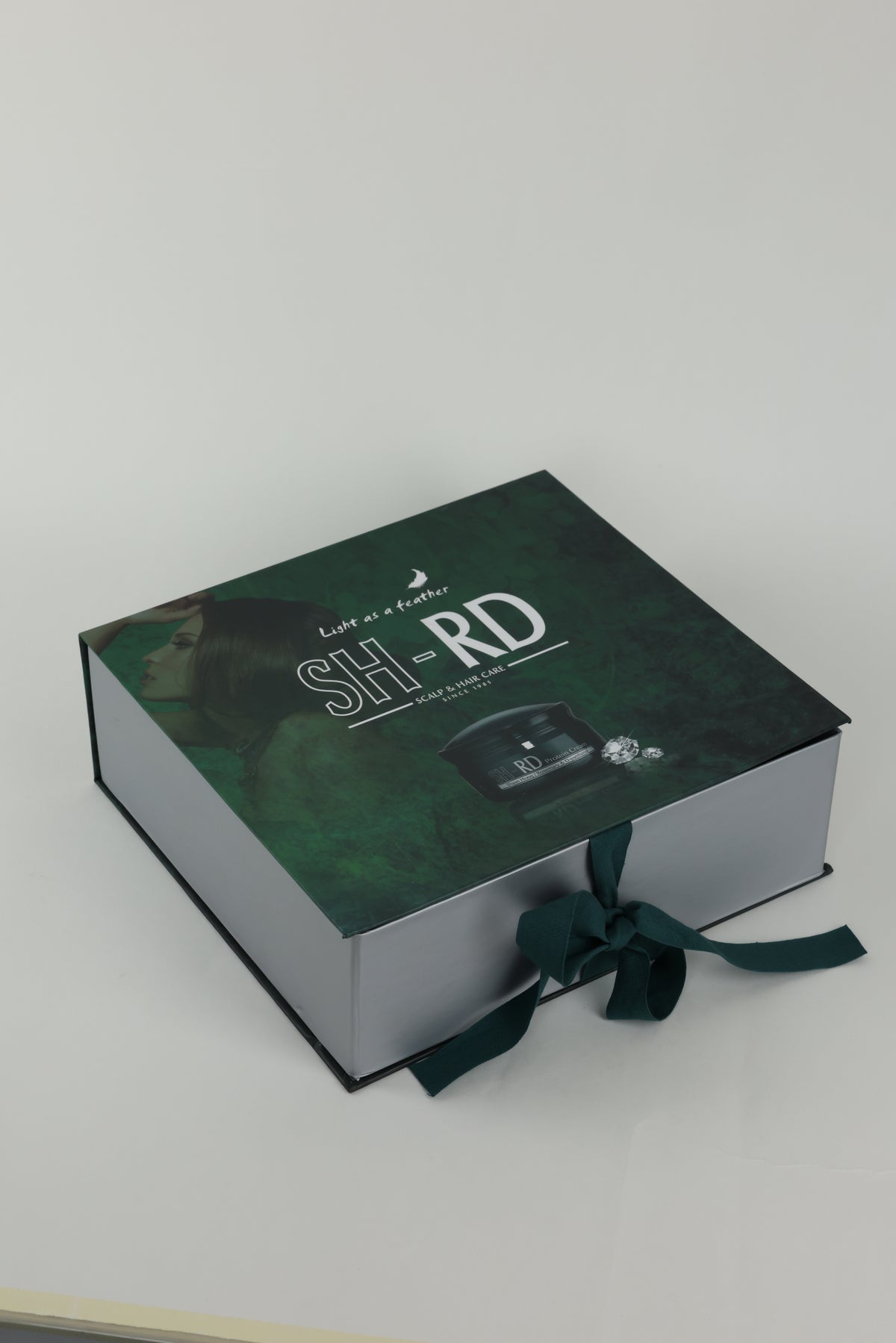 A rectangular gift box with a dark green textured lid and silver-gray sides, tied with a dark green ribbon. The lid features white text reading 'SH RD' and a small product photo, photographed on a light gray background.