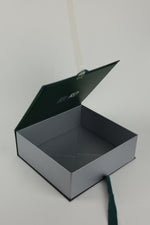 An open square presentation box with a dark green lid and a light gray interior, photographed on a plain light background.